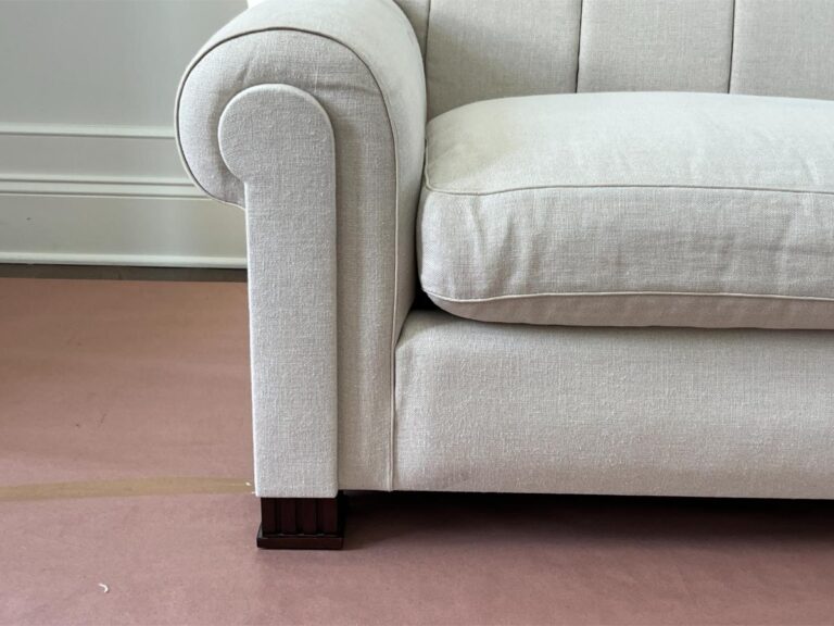 A close-up view of the armrest and seat cushion of a light beige sofa on a brown floor covering. The sofa has a rolled armrest and dark wooden legs.