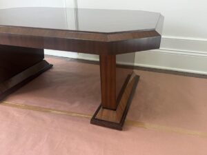 A close-up photo of a wooden table showing its angular leg and base design, positioned on a brown paper-covered floor.