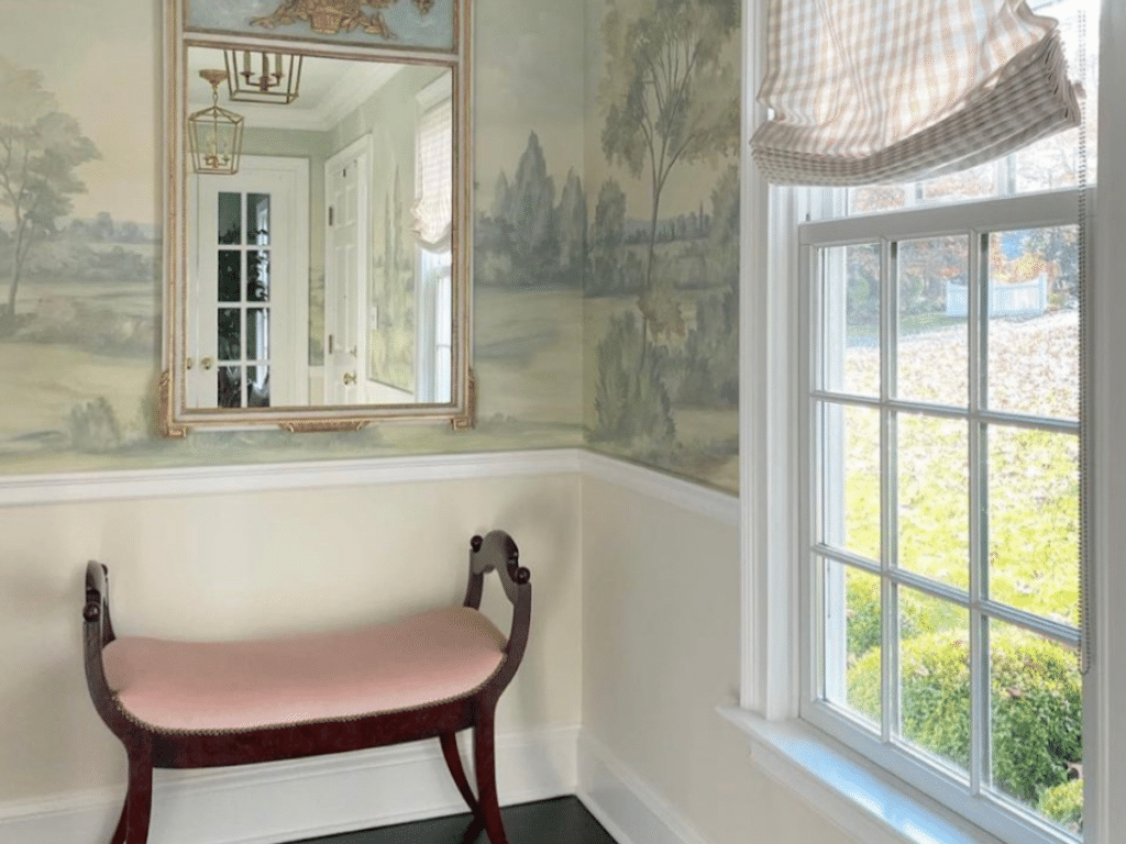 A small room with a landscape mural, a window with striped curtains, a pink-upholstered bench, a wall mirror, and a light fixture hanging from the ceiling.