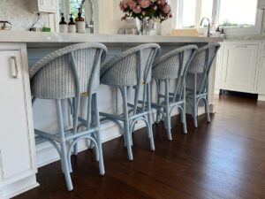 Four light gray Kitchen Stools by Loom USA are lined up at a white kitchen island, adorned with flowers and various kitchen items on the countertop. The kitchen showcases wooden floors and white cabinetry.