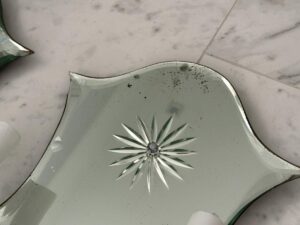 Close-up of a broken Vintage Mirrored Double Arm Sconce with a decorative star pattern, lying on a marble floor. The edges of the mirror are uneven and chipped.