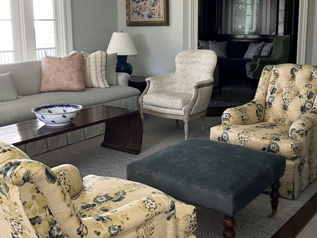 A living room with a beige sofa, two floral armchairs, a white armchair, a green ottoman, a wooden coffee table, and a blue and white bowl.