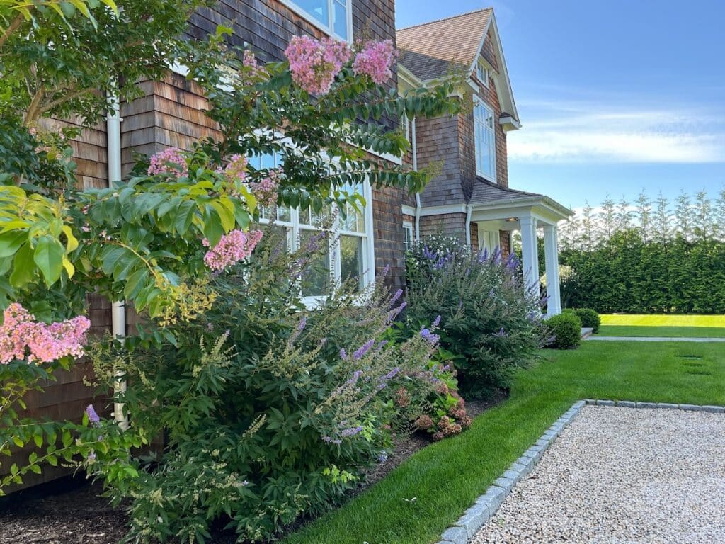 A landscaped garden with blooming flowers and shrubs adjacent to a wooden house, featuring a manicured lawn and gravel pathway under a clear blue sky.