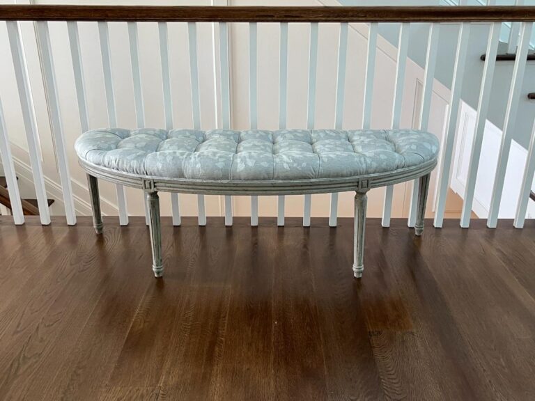 The Zentique Louis Curve Bench in a distressed olive finish, featuring a tufted cushion and four legs, is situated on polished hardwood flooring in front of white railings.