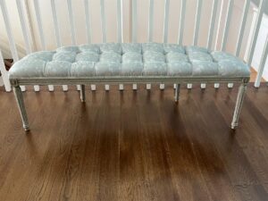 A Zentique Louis Curve Bench in Distressed Olive Finish with tufted upholstery and light blue fabric, featuring wooden legs, situated on a dark hardwood floor.