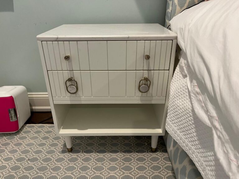 A white nightstand with two drawers and a shelf stands next to a bed. A small red and white speaker is on the floor beside the Niermann Weeks Tight Back Blue Sofas, Pair.