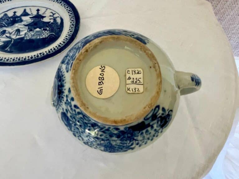 The bottom of a ceramic teapot with a blue and white pattern, marked with two labels reading "Gibbons" and "c1830, £275, K132".