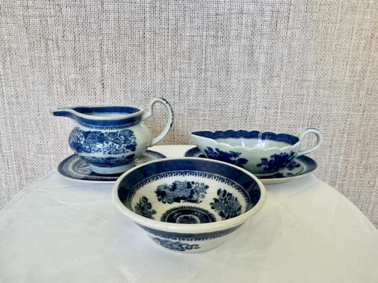 Three pieces of blue and white ceramic tableware, including a pitcher, a bowl, and a gravy boat, each resting on matching saucers against a textured beige backdrop.