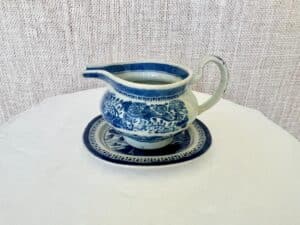 Blue and white ceramic pitcher on a matching saucer with intricate floral patterns, placed on a white tablecloth against a textured beige background.