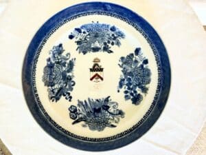 A round ceramic plate with a blue floral pattern and a central emblem featuring a shield and crown.