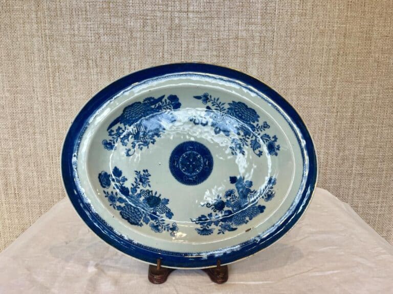 A decorative blue and white porcelain plate with floral and bird patterns, displayed on a wooden stand against a beige textured background.