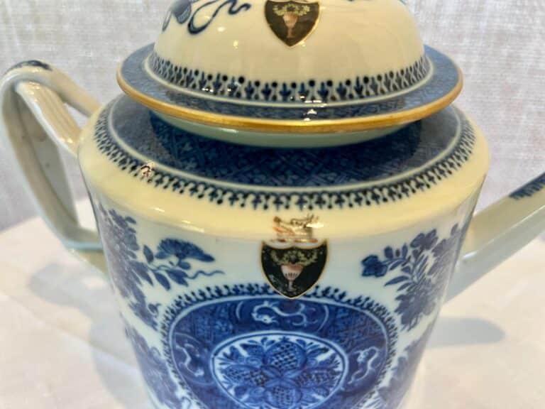 A blue and white porcelain teapot with intricate floral patterns and shield emblems on a light-colored background.