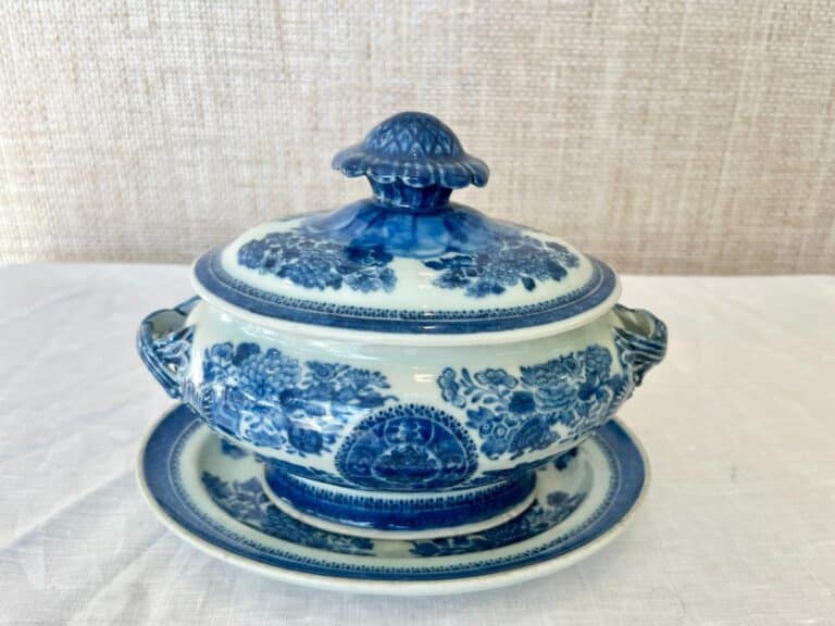 A blue and white ceramic soup tureen with floral patterns, featuring a lid, two handles, and a matching underplate, set on a white tablecloth.