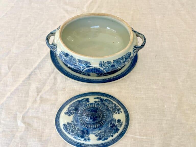 A blue and white ceramic soup tureen with floral patterns, featuring a lid and underplate, placed on a light fabric surface.