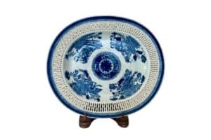 A round, blue and white porcelain antique Chinese Export Fitzhugh reticulated platter with intricate dragon patterns is displayed on a wooden stand.