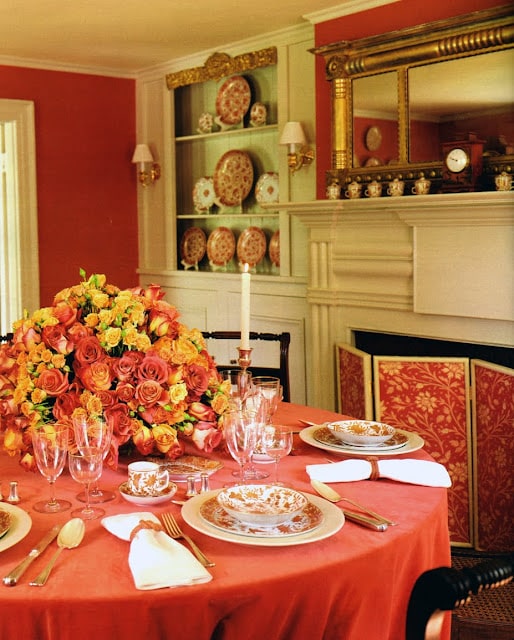 A formal dining room with an ornate table setting featuring a large bouquet of orange and yellow flowers. The room has a red and white color scheme, with a fireplace and decorative plates on display.