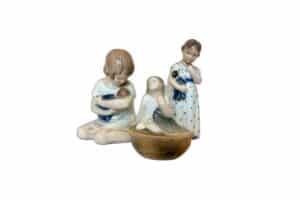 Three ceramic figurines of girls with brown hair, each holding a doll. Two are seated and one stands, all in light-colored dresses.