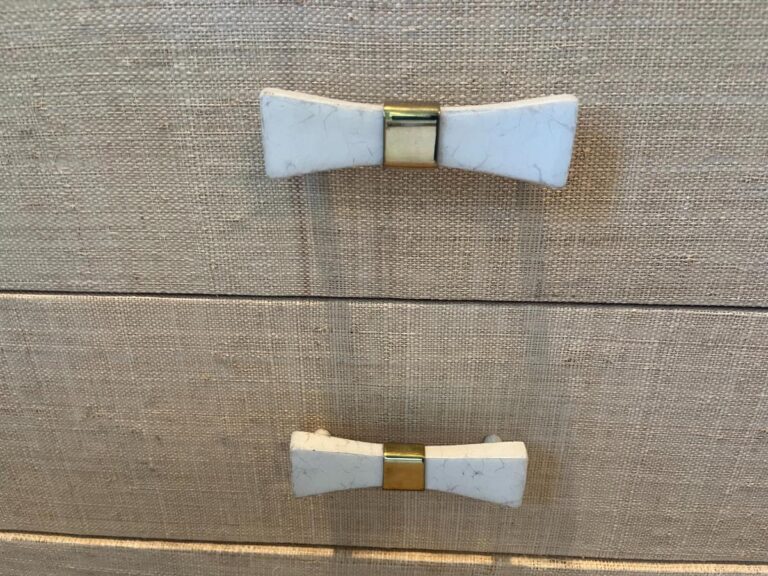 Two white and gold bow-shaped drawer handles resting on a pair of Niermann Weeks Tight Back Blue Sofas against a textured beige surface.