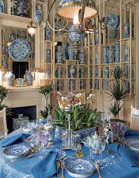 Elegant dining room with a blue-themed table setting, surrounded by shelves of decorative blue and white china, and adorned with plants and ornate lighting.