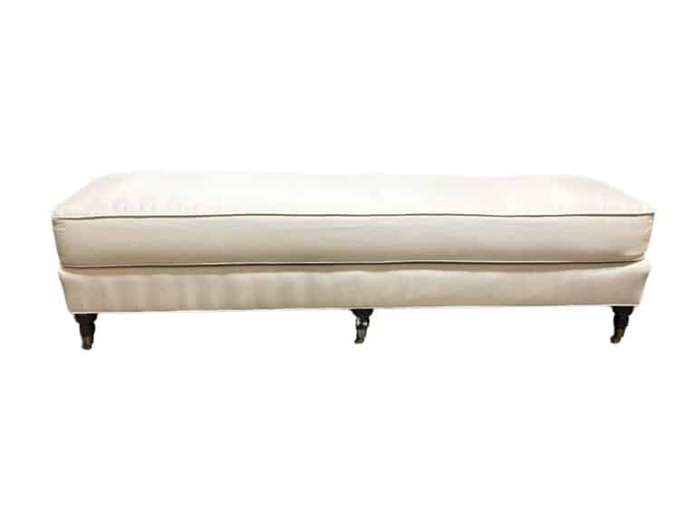 A long upholstered white bench with four dark wooden legs on casters.