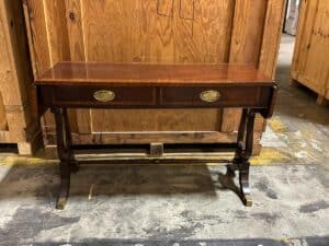 A wooden console table with two drawers and curved legs, set against a backdrop of wooden crates in a warehouse.