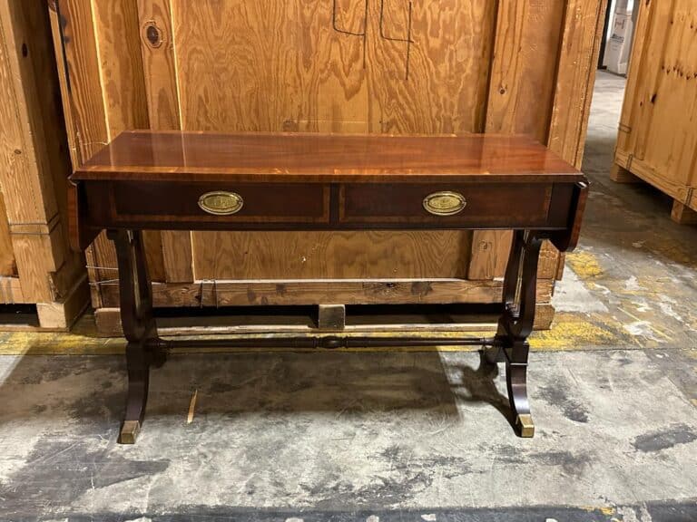 A wooden console table with two drawers and curved legs, set against a backdrop of wooden crates in a warehouse.