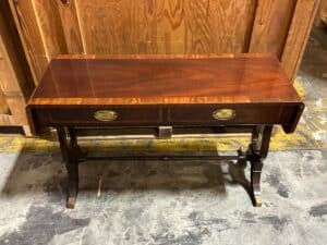 A wooden drop-leaf table with two drawers and brass handles stands on a concrete floor against a wooden background.