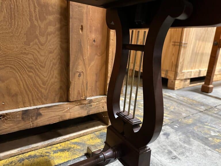 A wooden lyre-shaped table leg and crossbar, set against a backdrop of wooden furniture in a workshop.