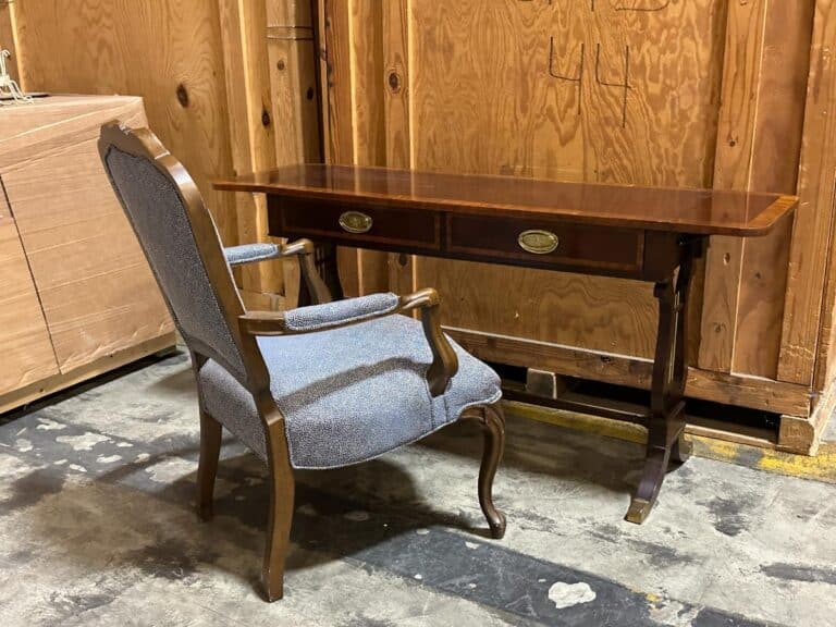 A wooden desk with two drawers and a cushioned chair are placed on a concrete floor in a storage area.