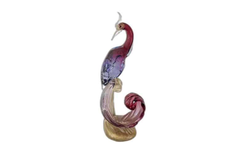 Stylized bird glass sculpture with a long tail, adorned in purple and red tones, elegantly perched on a golden base.