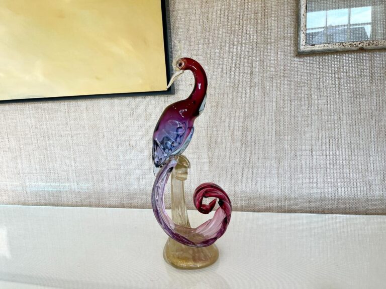 The Barbini Murano Bird of Paradise Italian Art Glass Sculpture features a bird with red and purple hues perched on a swirling base, beautifully displayed on a white surface with framed artwork in the background.