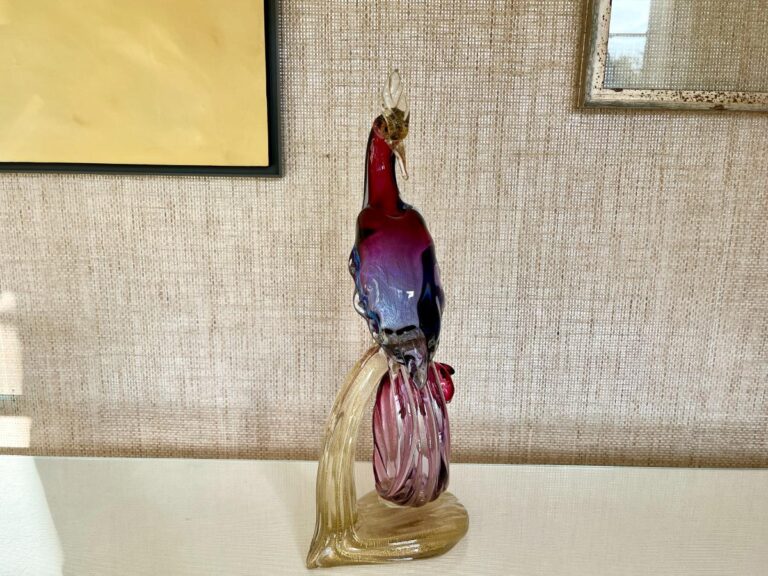 The Barbini Murano Bird of Paradise Italian Art Glass Sculpture, featuring a gradient red to blue body and clear tail, is showcased on a light-colored surface against a textured beige wall adorned with framed artworks.