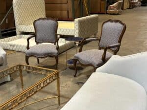 Two upholstered armchairs with wooden frames, a glass-top coffee table, a small round side table, and a cushioned bench in a warehouse setting.