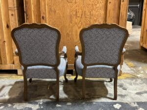 Two vintage armchairs with ornate wooden frames and blue upholstery are positioned back-to-back in a storage area with wooden crates.