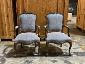 Two upholstered wooden chairs with ornate carvings are placed side by side on a warehouse floor, against a backdrop of wooden crates.