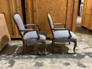 Two vintage upholstered chairs with wooden frames are placed back-to-back in a storage area with wooden crates and boxes.