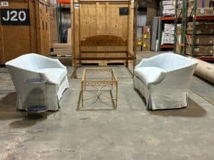 Two light blue sofas and a glass-top coffee table with a metal frame are arranged in a warehouse. A small round side table stands nearby. Background shows wooden crates and shelving.