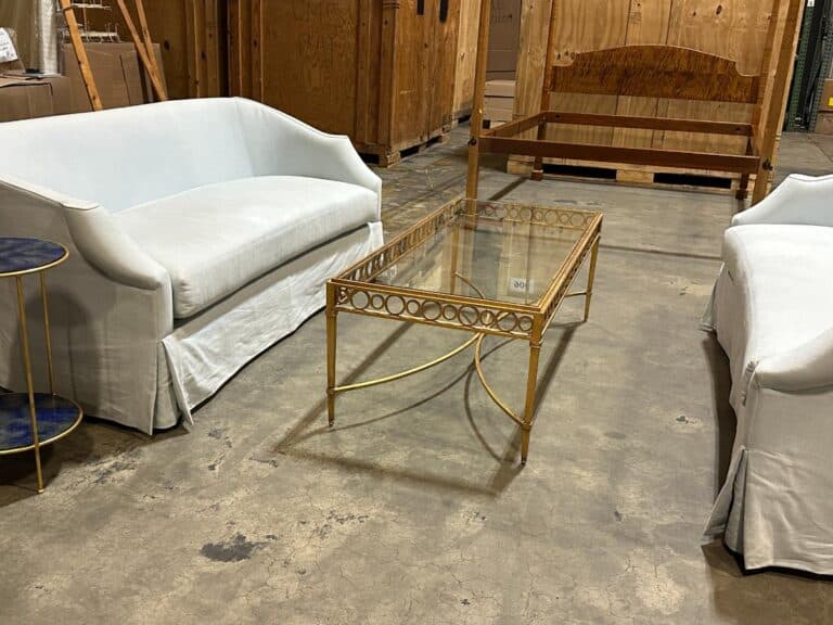 Two white sofas face each other with a glass coffee table in between. A round side table is next to one sofa. The setting is in a warehouse with wooden crates in the background.