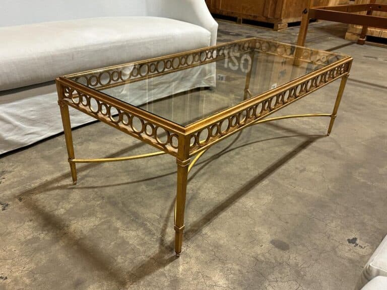 A glass-top coffee table with a gold metal frame featuring circular patterning, placed on a concrete floor near a white sofa.