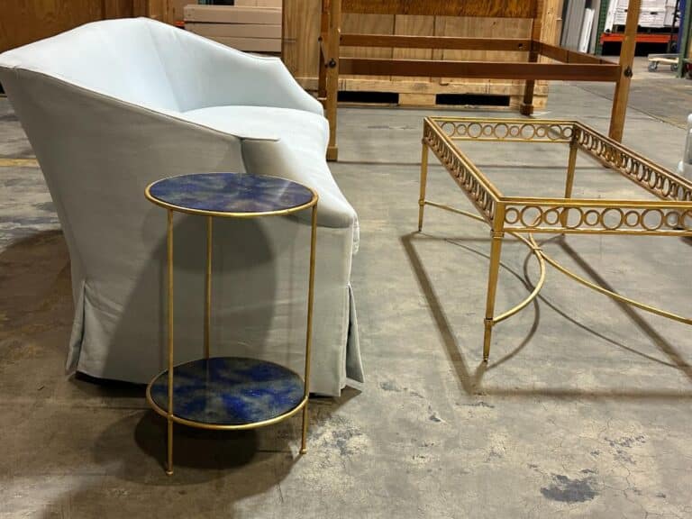 A light blue armchair next to a small round metal side table and a rectangular gold-framed glass coffee table on a concrete floor.