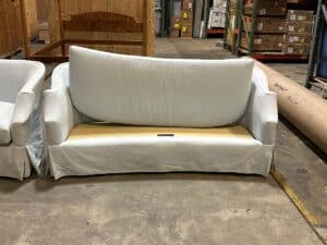 A white sofa with its back cushion removed, revealing a plain wooden panel, is displayed on a concrete floor in a warehouse setting.