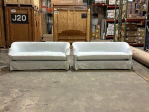 Two white upholstered couches in a warehouse setting with shelves, boxes, and wooden crates in the background.