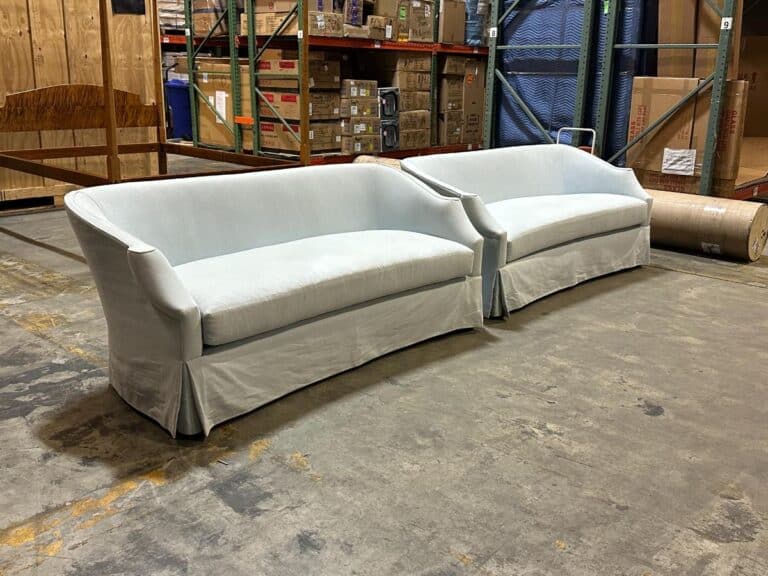 Two light gray sofas on a warehouse floor, surrounded by shelves with stacked boxes and packing materials.