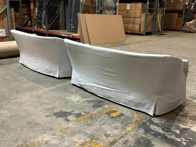 Two large, white-covered sofas placed back-to-back on a warehouse floor with cardboard boxes and metal shelves in the background.