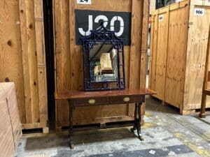 A wooden table with two drawers and an ornate mirror is placed in a storage area with large wooden crates in the background.