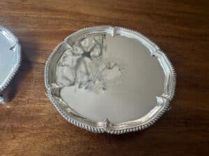 A round silver platter with ornate edges and an engraved design in the center, placed on a wooden surface.