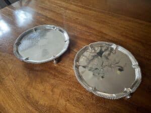 Two silver platters with ornate edges are placed on a wooden table.