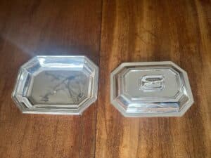 Two octagonal silver trays on a wooden surface. The tray on the left is uncovered, and the one on the right has a lid.