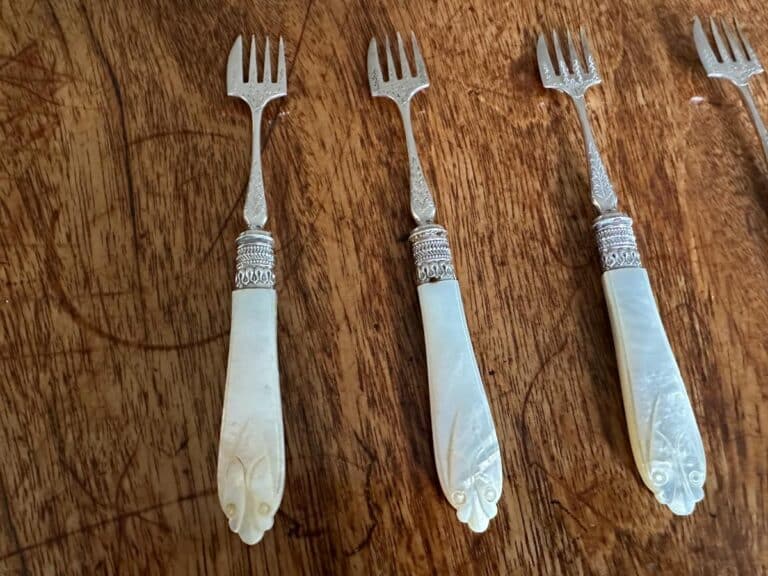 Three ornate seafood forks with engraved handles and mother-of-pearl grips are placed on a wooden surface.