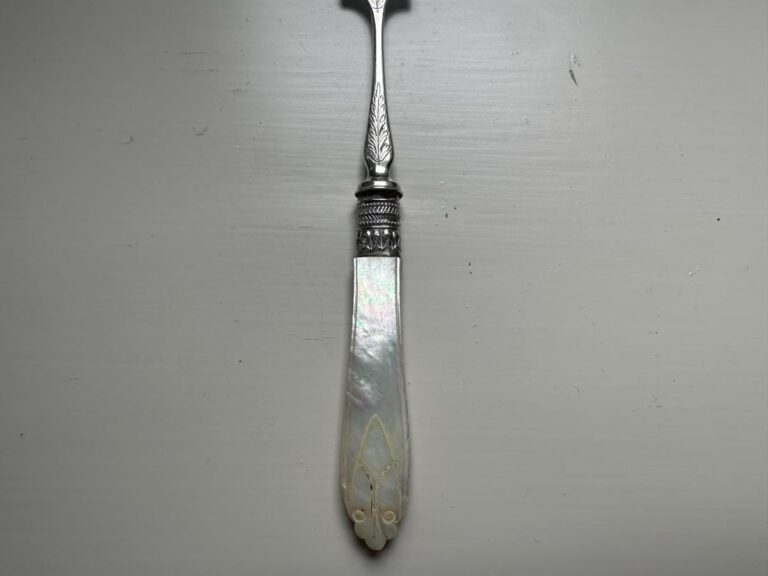 An ornate vintage butter knife with a decorative silver handle and a mother-of-pearl blade rests on a white surface.